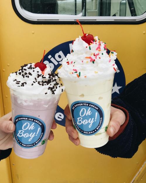Two colorful milkshakes topped with whipped cream, sprinkles, and cherries, held in front of a yellow ice cream truck.