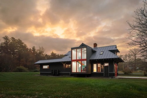 A modern black house with large windows, set against a dramatic sunset sky and surrounded by green grass and trees.
