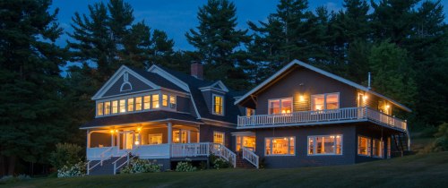 A beautifully lit two-story house surrounded by trees at dusk, featuring large windows and a welcoming porch.