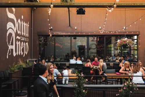 A lively outdoor dining area with string lights, people enjoying meals, and a sign reading "Saint Aneo."