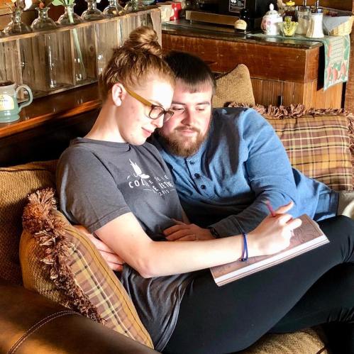 A couple sits together on a couch, sharing a sketchbook and smiling at each other in a cozy setting.