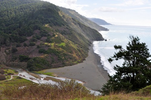 A scenic coastal view featuring a sandy beach, lush green hills, and a winding river flowing into the ocean.