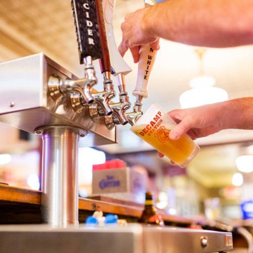 A hand pours a drink from a tap into a clear cup at a bar, with various beer taps visible in the background.