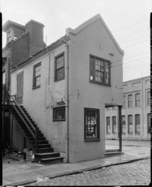 A small, two-story building with a staircase on the side, set in an urban environment with nearby structures.