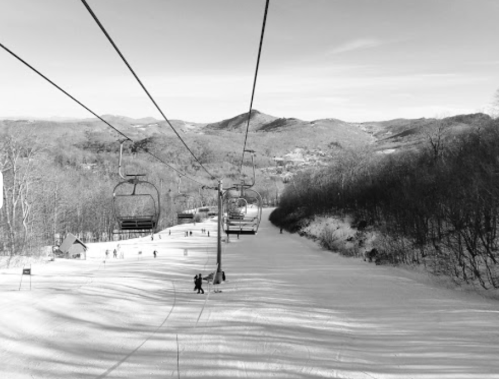A ski lift ascends a snowy slope, surrounded by trees and mountains under a clear sky.