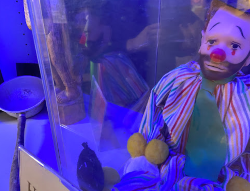 A clown doll in a display case, with colorful stripes and a sad expression, surrounded by blue lighting.