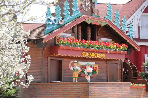 A charming wooden building with colorful flowers, featuring two cheerful figures in traditional attire and a water wheel.