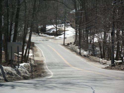 A winding road surrounded by trees, with patches of snow and a sign indicating the road number.