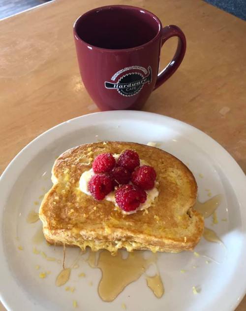 A plate of French toast topped with raspberries and syrup, next to a coffee mug on a wooden table.