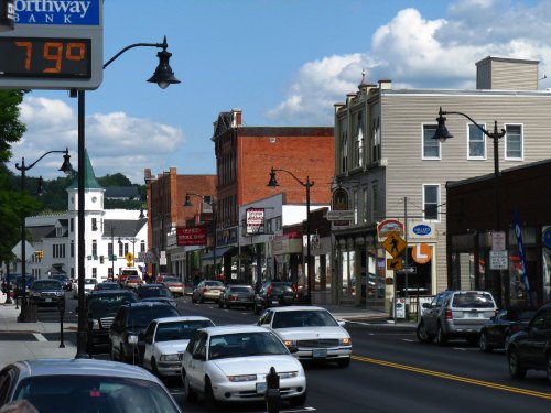 A busy street scene with shops, cars, and a bank sign, featuring a church steeple in the background under a blue sky.