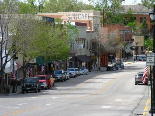A quaint street lined with shops and trees, featuring parked cars and American flags, in a small town setting.