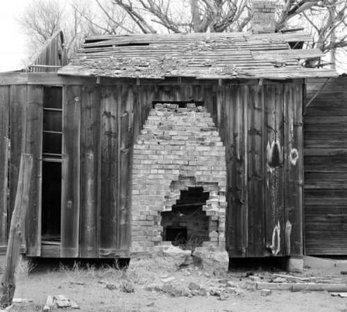 A weathered wooden building with a crumbling brick chimney and a partially collapsed roof, set in a barren landscape.