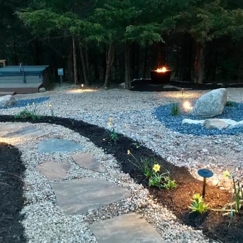A serene garden path with stone walkways, flowers, and soft lighting, leading to a fire pit surrounded by trees.