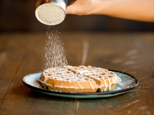 A hand sprinkles powdered sugar over a freshly made waffle on a plate.