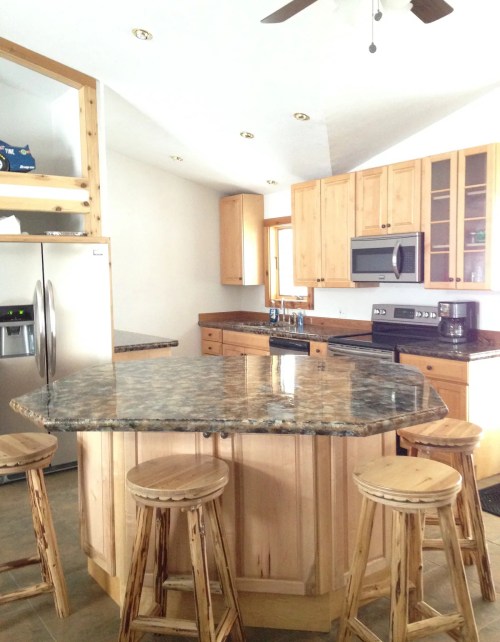 A modern kitchen featuring wooden cabinets, a granite island, and three wooden stools. Bright and spacious design.