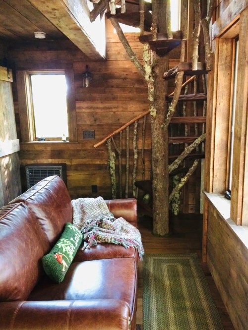 Cozy interior of a rustic cabin featuring a leather sofa, a woven blanket, and a spiral staircase made of tree branches.