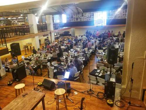 A large gathering of people seated at tables in a spacious venue, with a stage and sound equipment in the foreground.