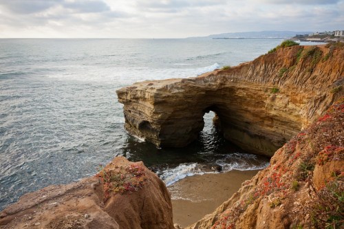 A rocky coastal arch with waves crashing below, surrounded by cliffs and sandy beach under a cloudy sky.