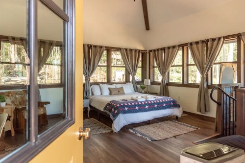 Cozy bedroom with a large bed, natural light from windows, and rustic decor, surrounded by trees.
