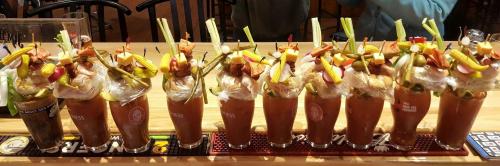 A row of colorful, garnished Bloody Mary cocktails on a bar, featuring celery, olives, and various fruits.