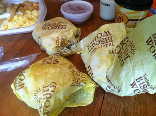 Three wrapped breakfast sandwiches from Tudor's Biscuit World on a wooden table, with a side of scrambled eggs and a drink.
