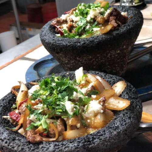 A stone bowl filled with sizzling meat, onions, and herbs, topped with crumbled cheese.