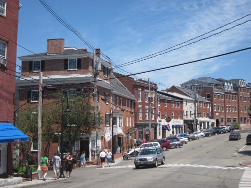 A sunny street scene with brick buildings, shops, and people walking along the sidewalk under a blue sky.