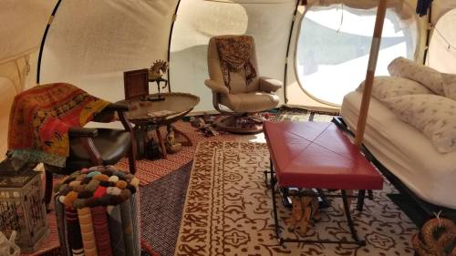 Cozy interior of a tent featuring a chair, table, patterned rugs, and a bed with pillows. Warm, inviting atmosphere.