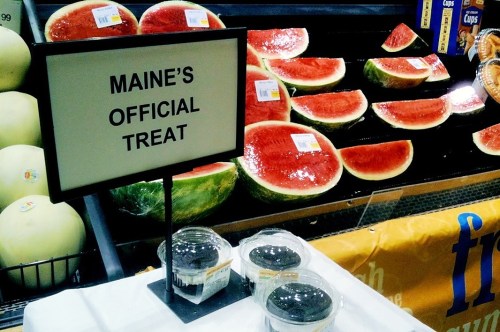 A display of watermelons with a sign reading "Maine's Official Treat" in a grocery store.