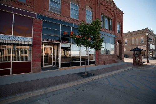 Exterior view of a coffee shop with large windows, a small tree, and a historic brick building in a quiet street.
