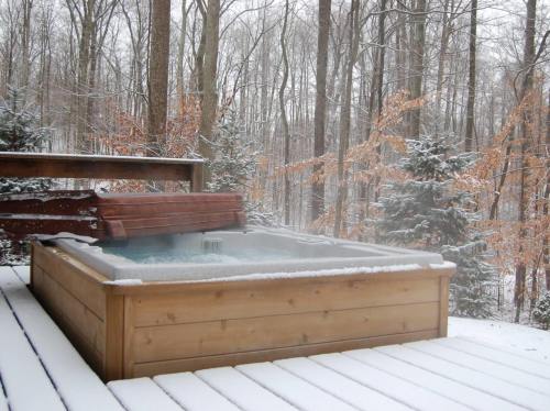 A wooden hot tub on a snowy deck, surrounded by trees in a winter landscape. Steam rises from the water.