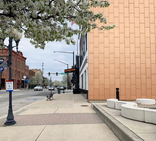 A street view showing a modern building with a copper facade, flowering trees, and a bench on a sidewalk.