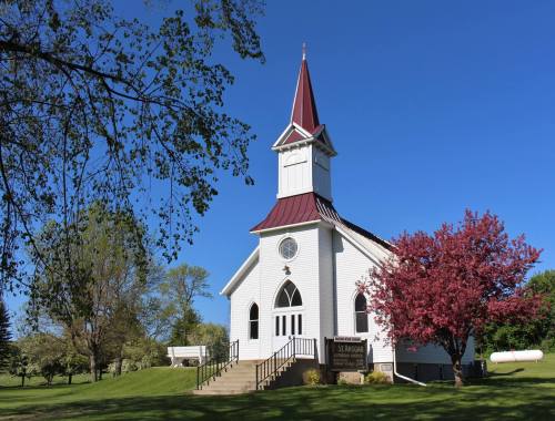 A white church with a red steeple, surrounded by green trees and a blooming pink tree under a clear blue sky.