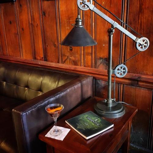 A cozy corner with a leather couch, a cocktail on a table, and a vintage lamp illuminating a book.