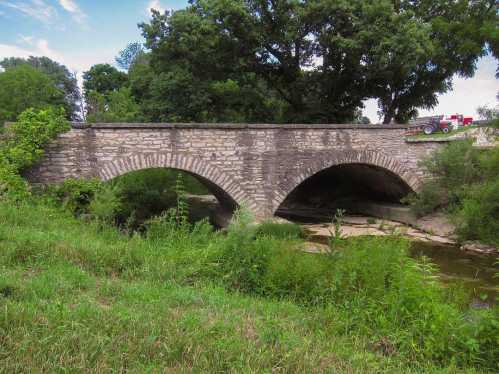 A stone arch bridge spans a small river, surrounded by lush greenery and trees under a blue sky.