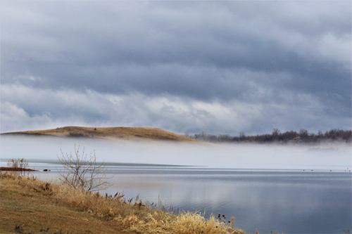 A serene lake scene with fog rolling over the water, surrounded by grassy banks and distant hills under a cloudy sky.