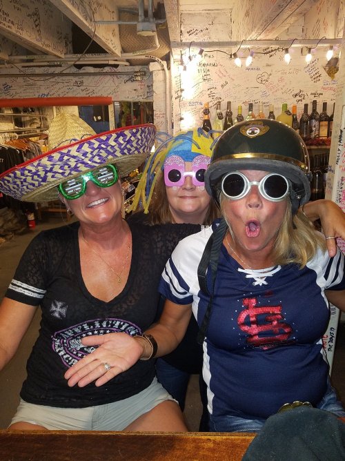 Three women wearing fun hats and oversized sunglasses, posing playfully in a lively bar setting.