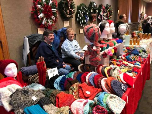 Two women sit behind a table filled with colorful knitted hats and scarves, surrounded by festive wreaths.