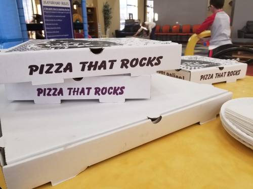 Three pizza boxes labeled "Pizza That Rocks" on a table, with plates and a blurred background of a playground area.