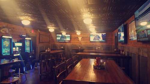 A cozy bar interior with wooden tables, slot machines, and multiple TVs displaying sports, illuminated by warm lighting.