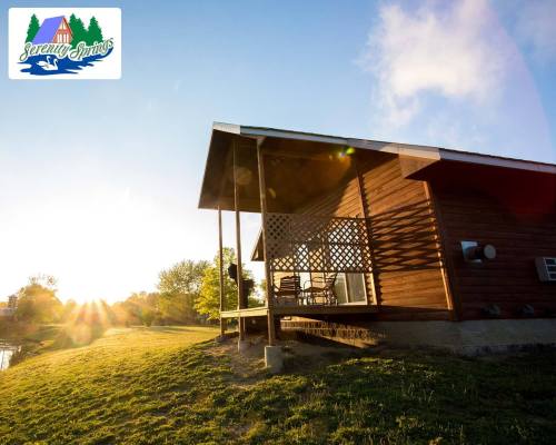 A cozy cabin at sunset, with a porch and chairs, surrounded by greenery and a peaceful landscape.