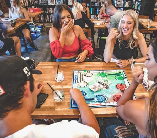 A group of friends laughing and playing a board game at a lively café, with drinks on the table.