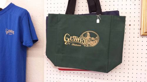 A green tote bag with "Guthrie, Oklahoma" printed in gold, displayed alongside a blue t-shirt on a pegboard.