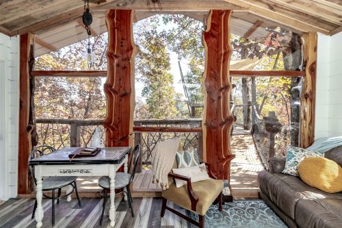 Cozy cabin interior with wooden beams, a small table, and a view of trees through large windows.