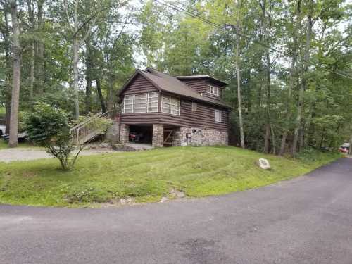 A rustic wooden house with a stone foundation, surrounded by trees and a grassy area, located near a winding road.