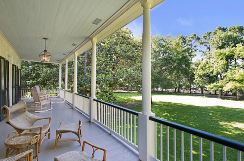 A spacious porch with rocking chairs overlooks a lush green yard and trees under a clear blue sky.