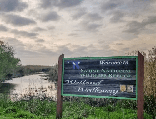 Sign welcoming visitors to Sabine National Wildlife Refuge, featuring a wetland walkway and a cloudy sky.