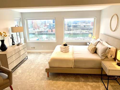 A cozy bedroom with a large bed, decorative pillows, and windows showcasing a city view. Soft lighting and modern decor.