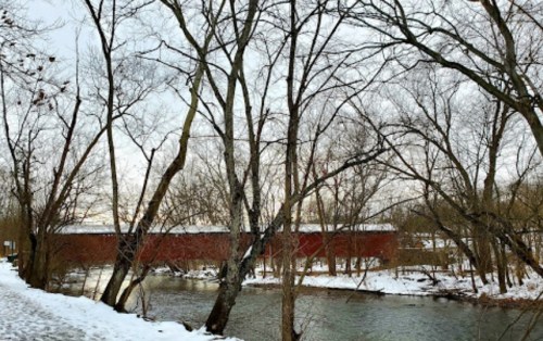 A snowy landscape featuring bare trees along a river, with a red covered bridge in the background.