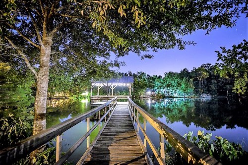 A wooden dock extends into a calm lake, surrounded by trees, under a starry night sky with soft lighting.